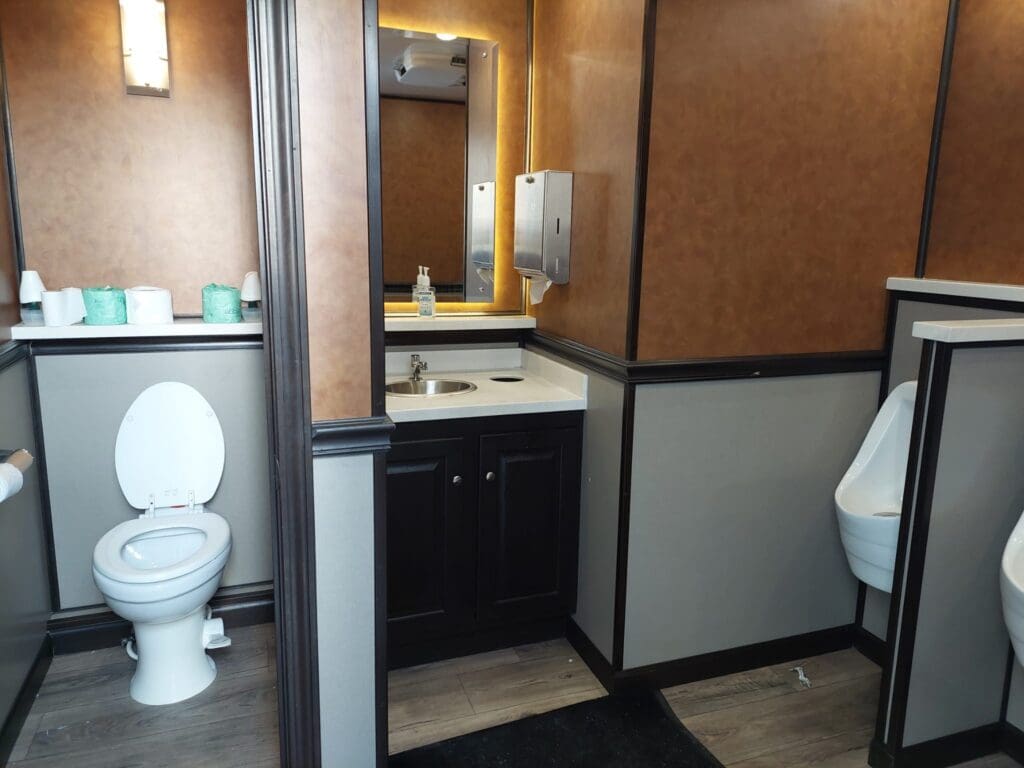 Warm wood and copper-tone restroom trailer interior with vanity, mirror and stalls