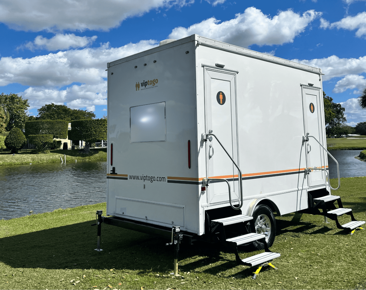VIP To Go luxury restroom trailer set up on a lawn beside a pond under a blue sky